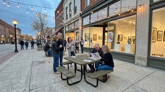 A group of people sitting at a gold picnic table on the sidewalk outside an art gallery 