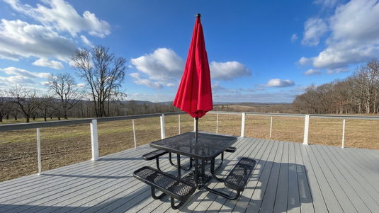 a black picnic table with a red umbrella on a porch with cloudy blue skies and rolling hills in the background