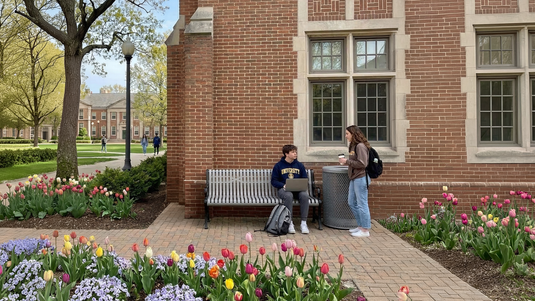 two college kids talking on a grey bench in early spring on campus