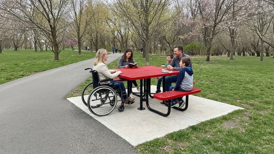 A family sits at a red ADA accessible picnic table in a park with budding trees on a sunny spring day