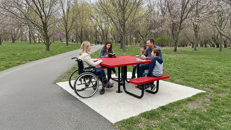 A family sits at a red ADA accessible picnic table in a park with budding trees on a sunny spring day