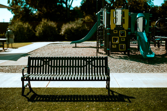 Park seating area with a black bench facing a playground surrounded by trees and landscaping.