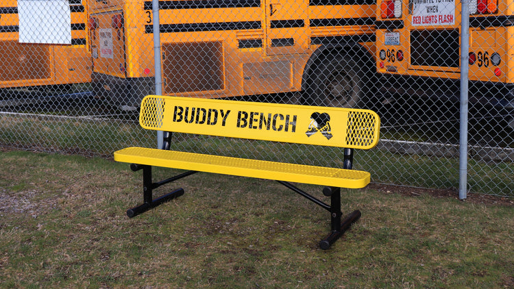 A yellow buddy bench with school busses behind it