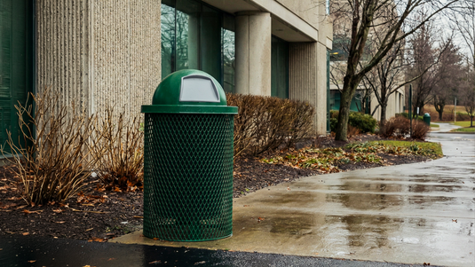 A green trash can outside of an office building on a rainy day