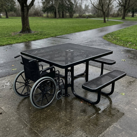 Wheelchair next to a black metal ADA accessible picnic table on a wet pavement with grass and trees in the background.