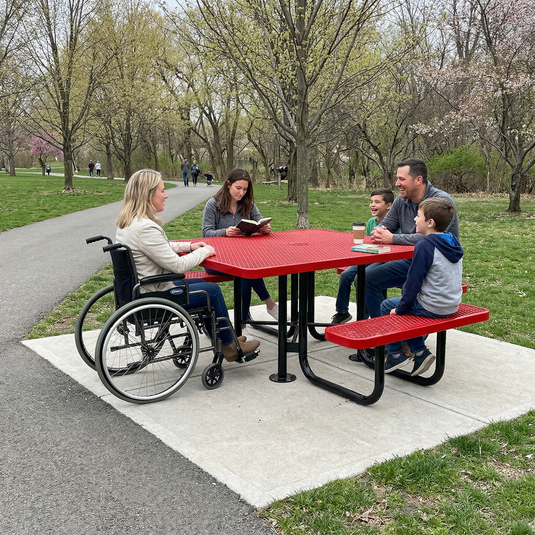 Family with a person in a wheelchair at a red ADA accessible picnic table in a park.