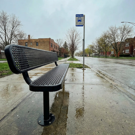 a black park bench with raindrops on it sat beside a bus stop sign in a neighborhood on a rainy day