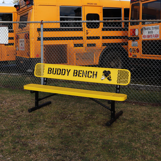 Yellow 'Buddy Bench' in front of a chain-link fence with school buses in the background.